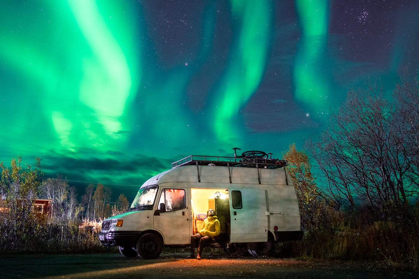 man sat on step of campervan looking up at night sky with northern lights